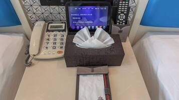 A hotel room with a bedside telephone, a digital clock, a tissue box, and a notepad on the bed.