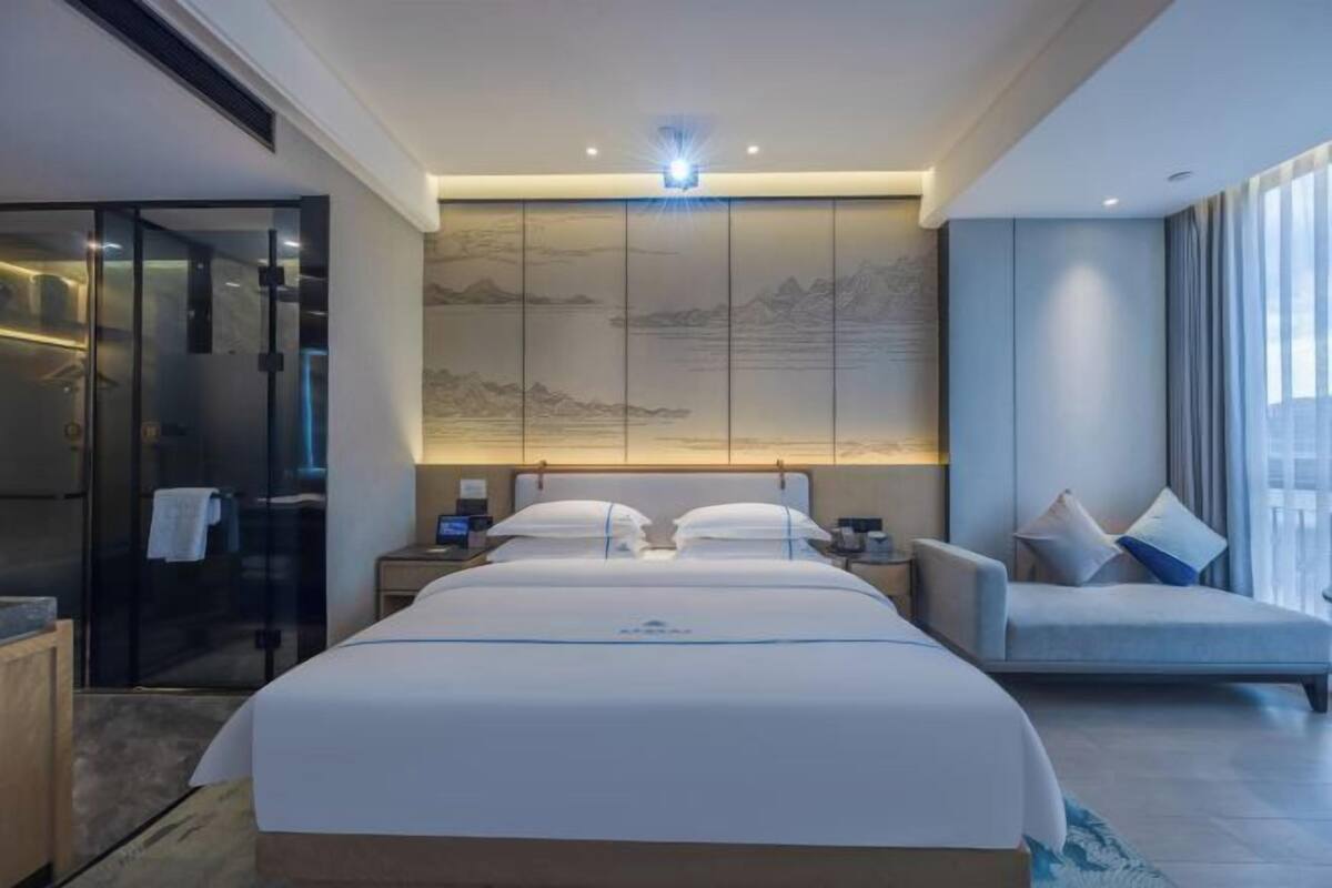 Una habitación de hotel moderna con cama, zona de estar, cabina de ducha de vidrio y un mural de paisaje montañoso en la pared.
