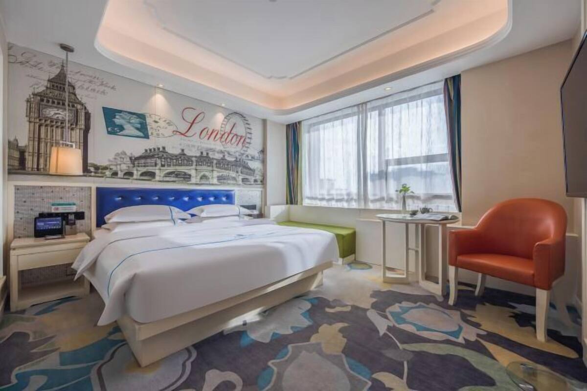 Una habitación de hotel con una cama, un escritorio con computadora, una silla, un ventanal con cortinas y una pared con un mural de paisaje urbano.