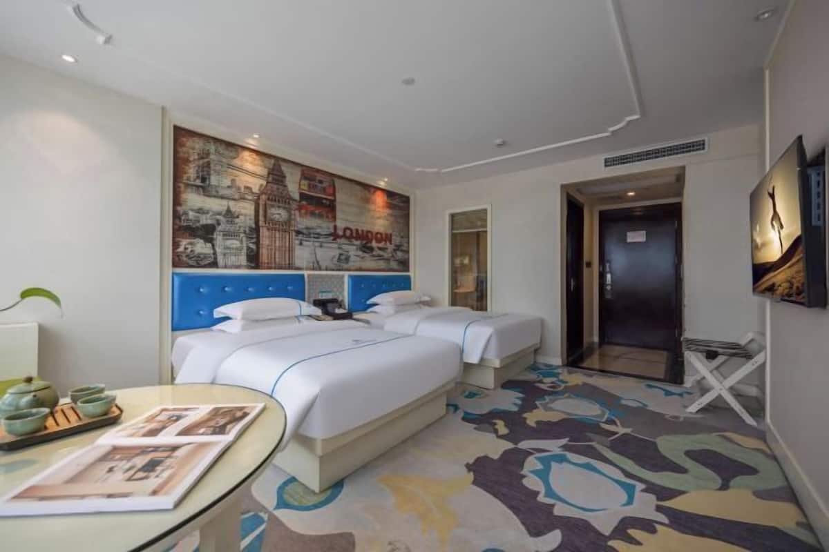 Una habitación de hotel con dos camas, un cartel grande con un paisaje urbano, una mesita de vidrio con revistas y una alfombra estampada.