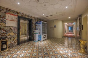 Ein Hotelkorridor mit gemustertem Boden, einem Verkaufsautomaten und einem Spiegel an der linken Wand.