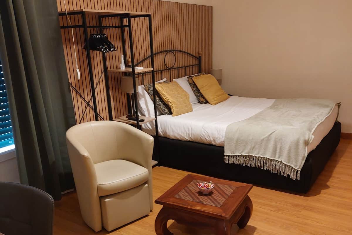 A hotel room with a bed, a chair, a small wooden table, and a window with curtains.