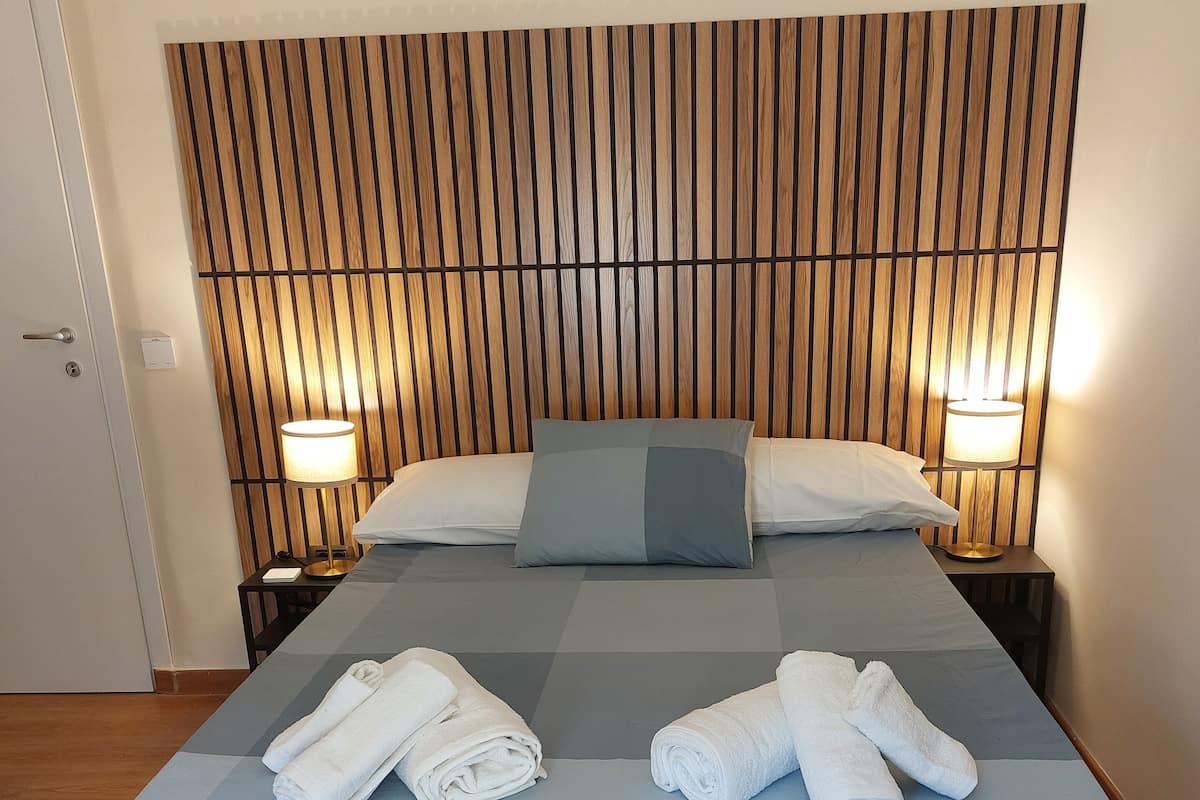 A neatly made bed with a blue mattress and two pillows, a wooden headboard, two bedside lamps, and two rolled towels on the bed.
