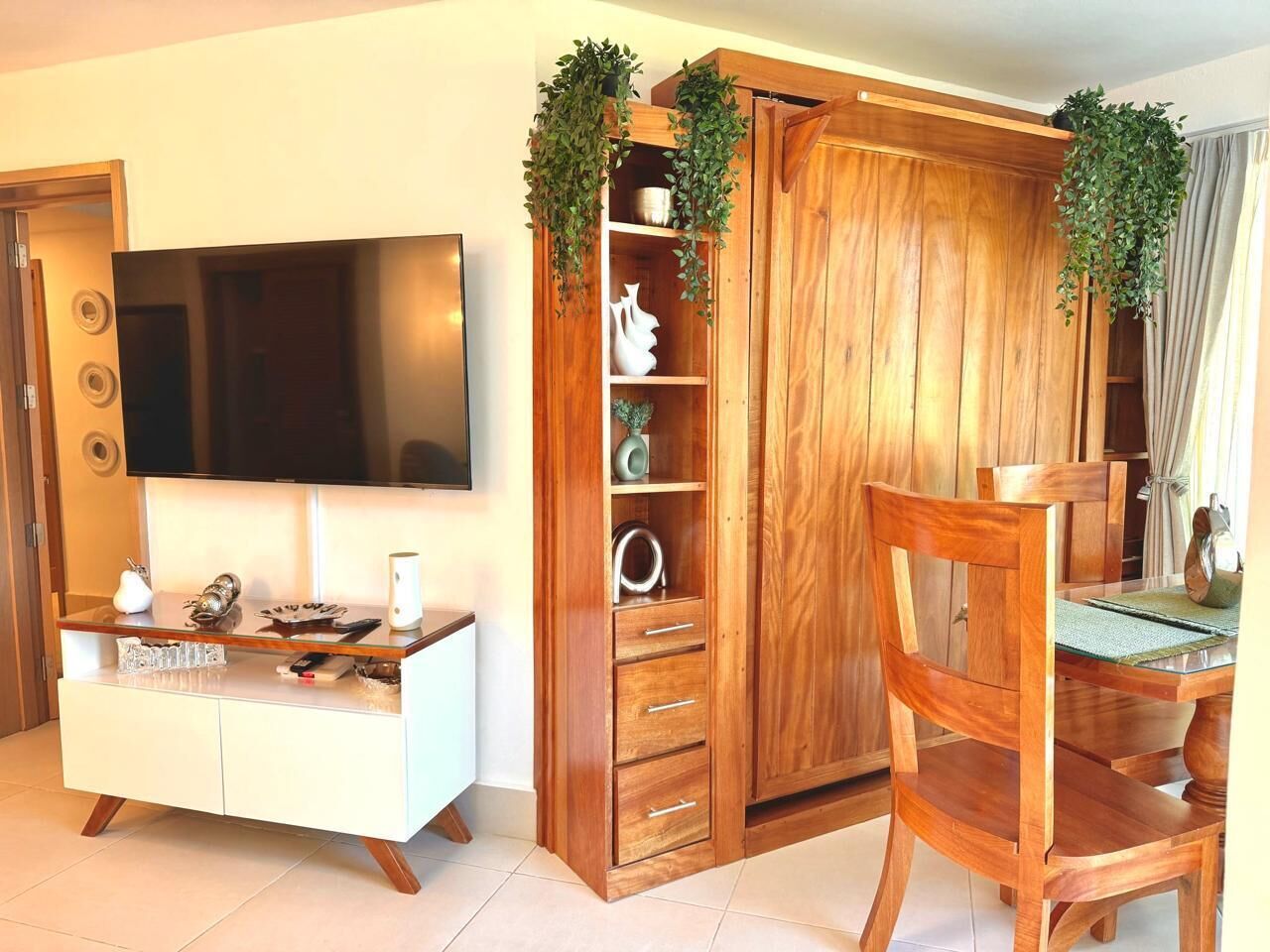 Una sala de estar moderna con un televisor de pantalla plana, un mueble de madera con estantes, y un comedor con una mesa y sillas de madera.