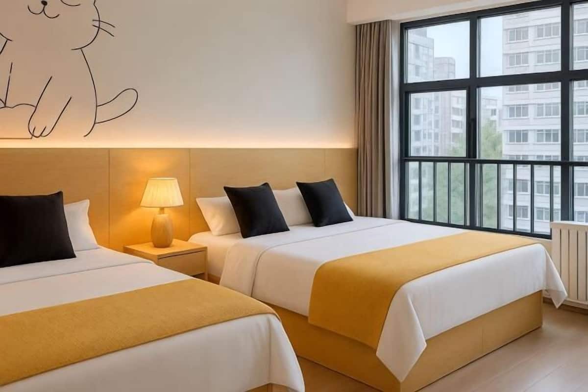 A hotel room with two beds, a nightstand with a lamp, a large window with a view of buildings, and a wall art of a cat.
