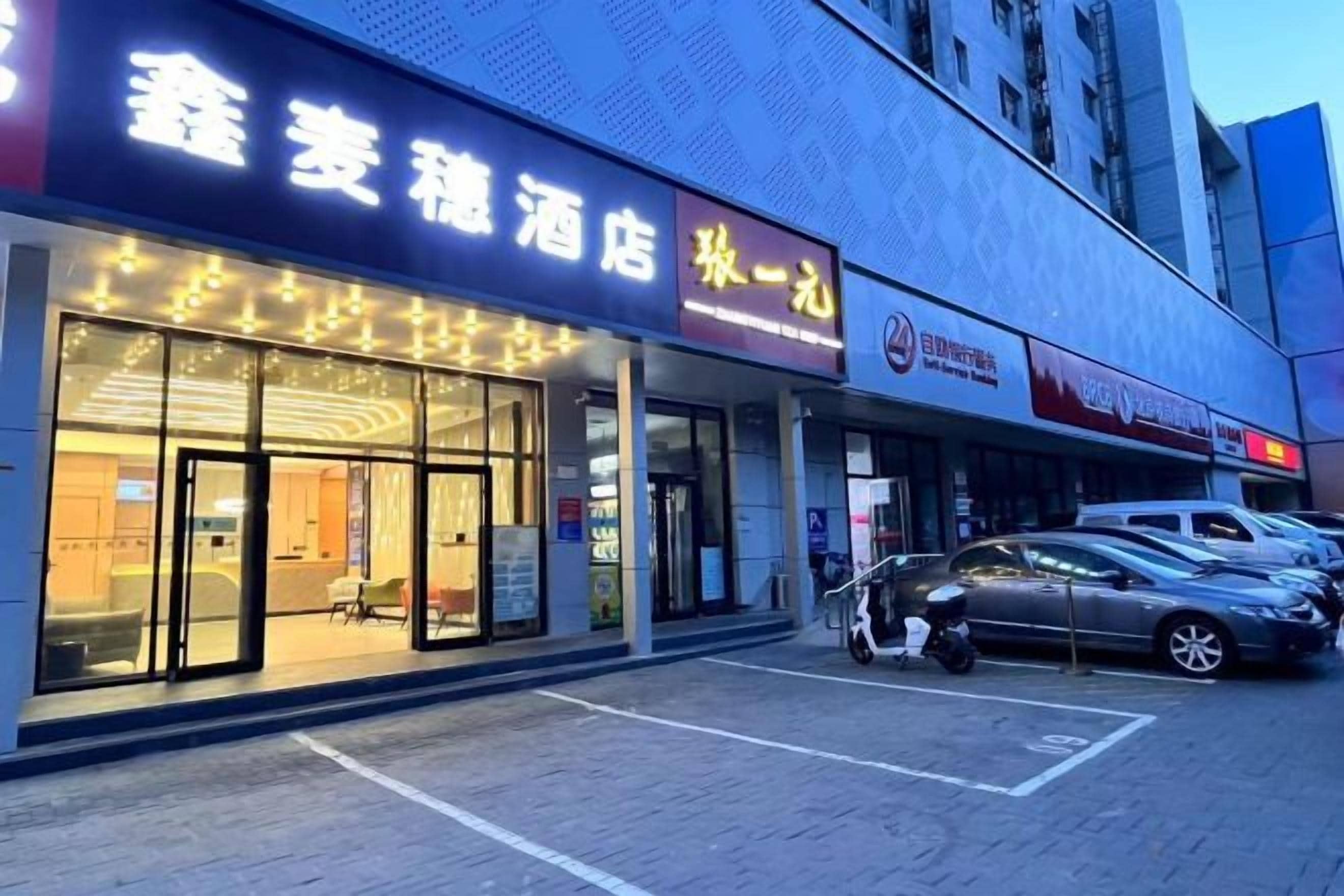 Hotel con fachada de vidrio, entrada visible y estacionamiento con automóviles.