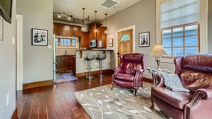 A spacious living room with a leather sofa, a red leather chair, and a bar area with stools. There is a large window with blinds, a ceiling fan, and a kitchen in the background. - Cozy Heights Hideaway :Pets welcome-Big Frontyard (Houston)