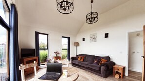 A spacious living room with a dark grey sofa, a round wooden coffee table, a laptop, and a TV on a wooden stand. There are large windows, a ceiling light fixture, and a door to the right. - The Flower Room - sleeps 2 guests  in 1 bedroom (Droitwich)