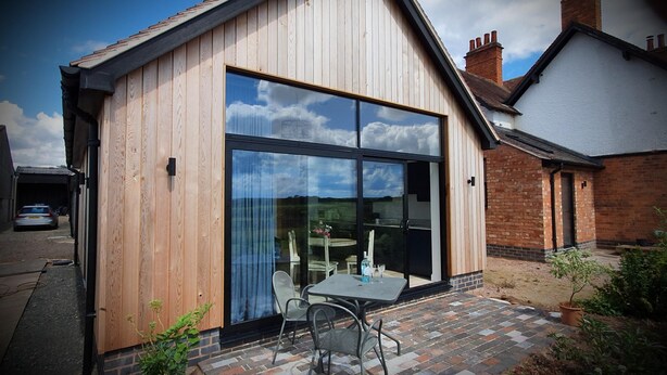 A modern wooden house with large glass windows, a patio with a table and chairs, and a brick pathway. - The Flower Room - sleeps 2 guests  in 1 bedroom (Droitwich)