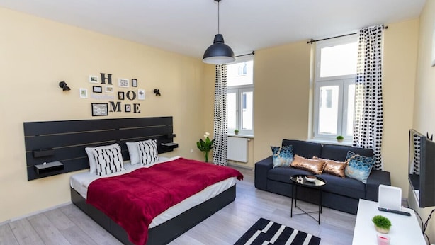 A modern bedroom with a bed, a sofa, a desk, and a TV. - The Glowing Ruby | By Radiant Apartments (Wien)