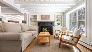 Una sala de estar con sofá, sillón, mesa de centro de madera, chimenea y ventanas grandes.