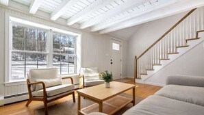 Un salón amplio con suelo de madera, paredes blancas, un ventanal con vistas a un paisaje nevado, una escalera y un sofá blanco.