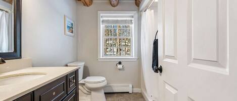 Een badkamer met een wit toilet, een wastafel met spiegel, een raam met uitzicht op bomen, en een deur met een zwarte hendel.