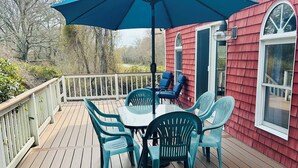 A wooden deck with a table and chairs under a blue umbrella, a red house in the background, and a white railing.