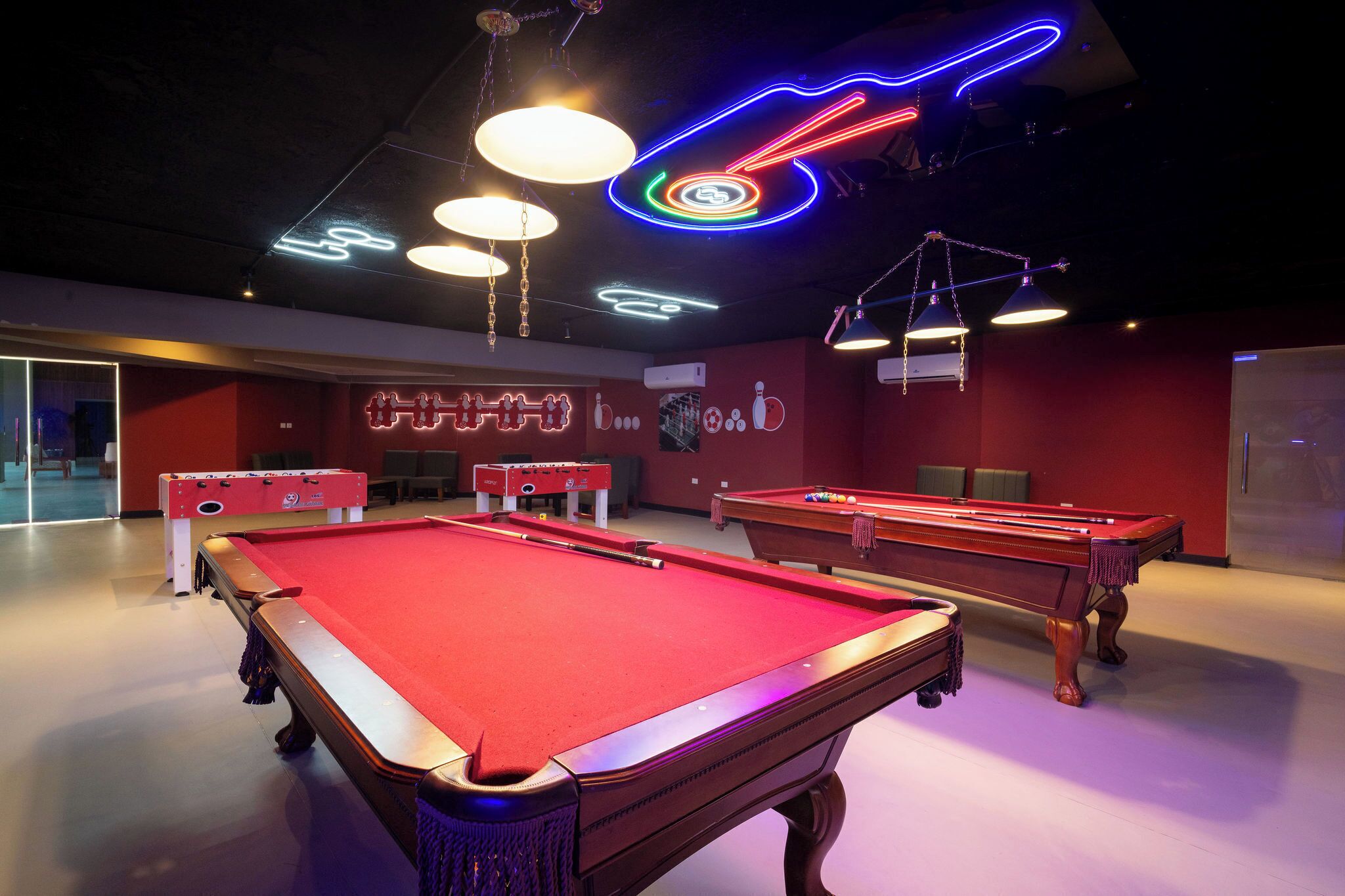 A pool table with a red felt surface, a foosball table, and neon signs on the ceiling.