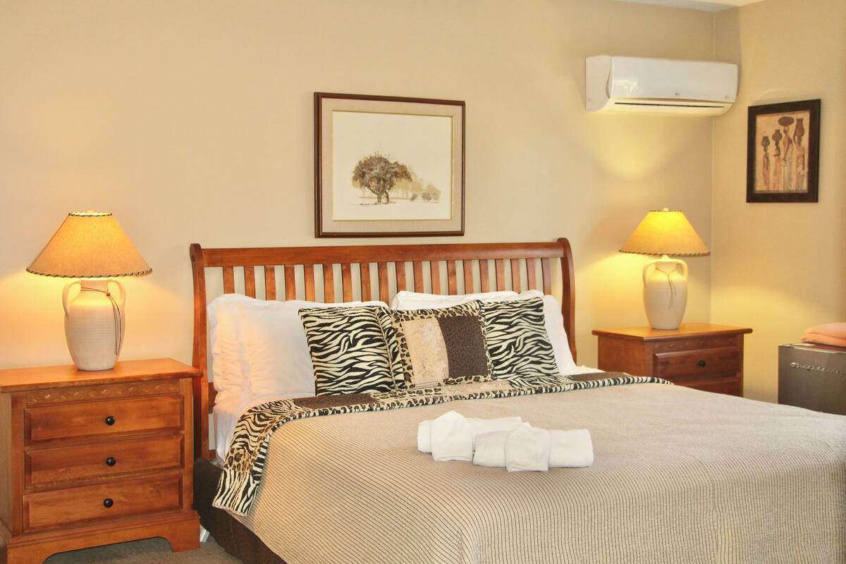 Een net opgemaakt bed met kussens in zebraprint, een nachtkastje met een lamp en een lijstje met een foto aan de muur.