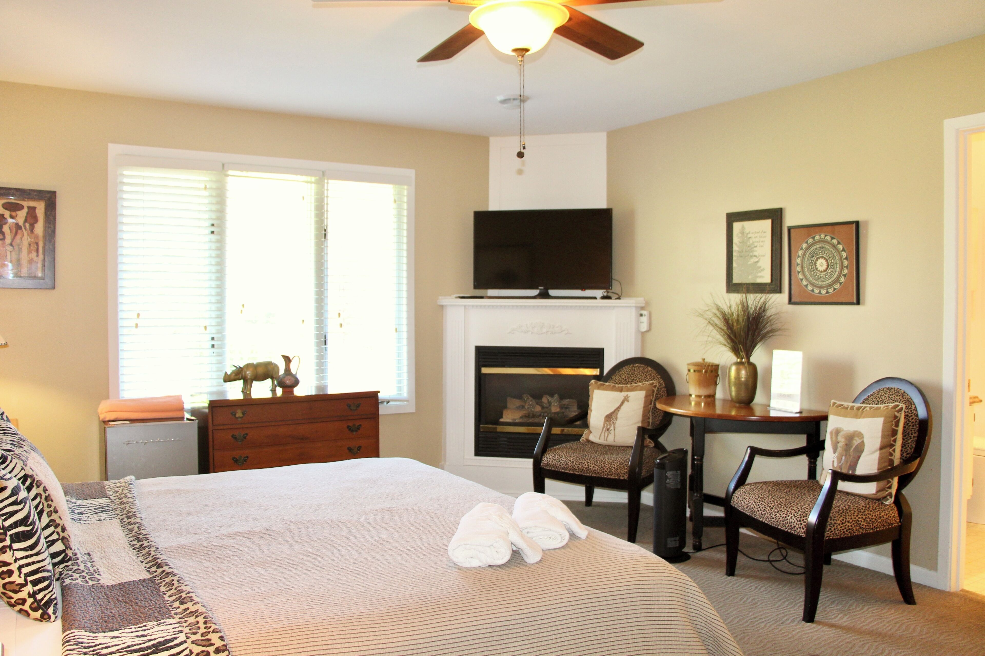 A bedroom with a bed, two chairs, a fireplace, a television, and a ceiling fan.