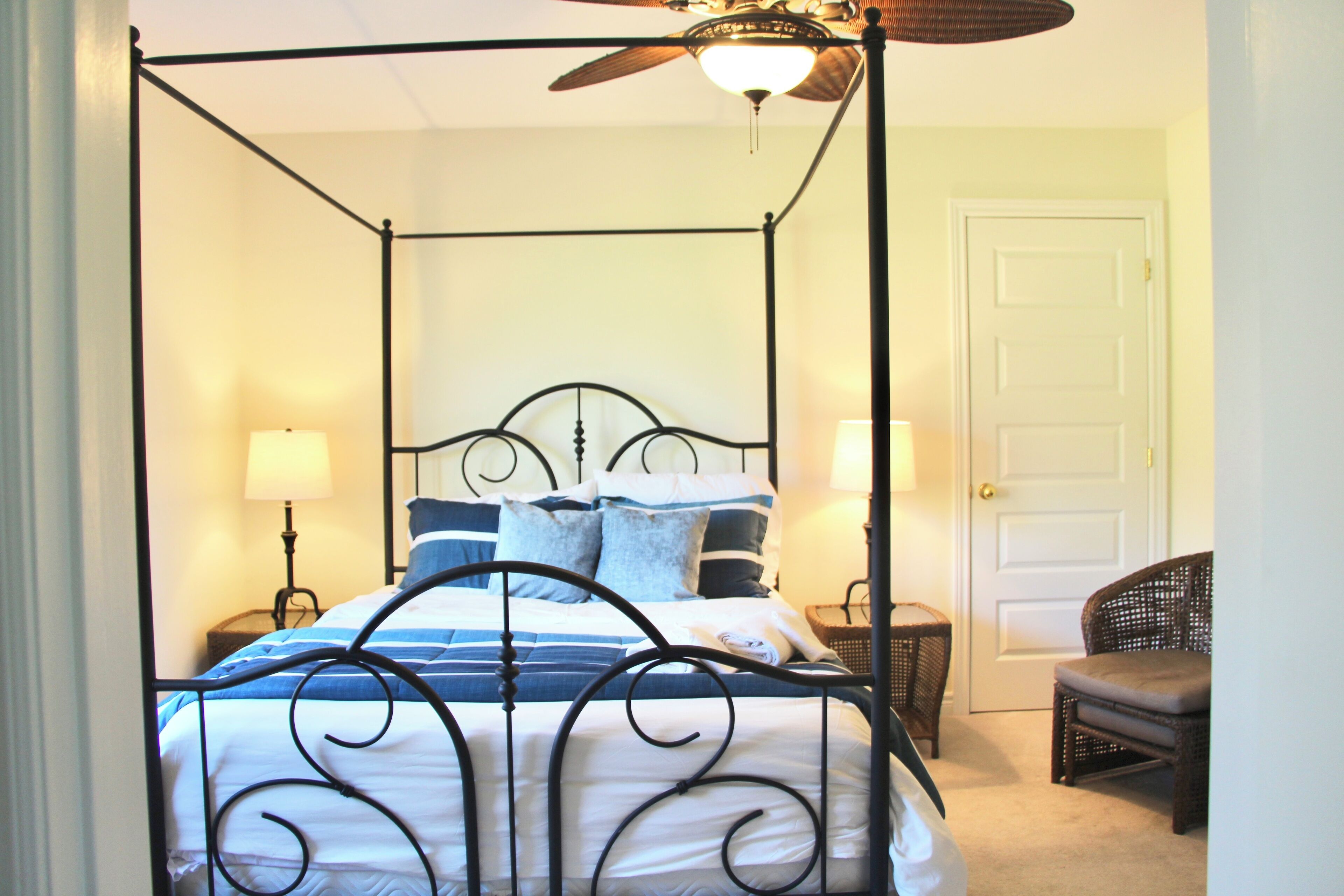 A four-poster bed with a ceiling fan, a wicker chair, and a door in a room with a carpeted floor.