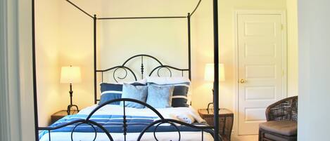A four-poster bed with a ceiling fan, a wicker chair, and a door in a room with a carpeted floor.