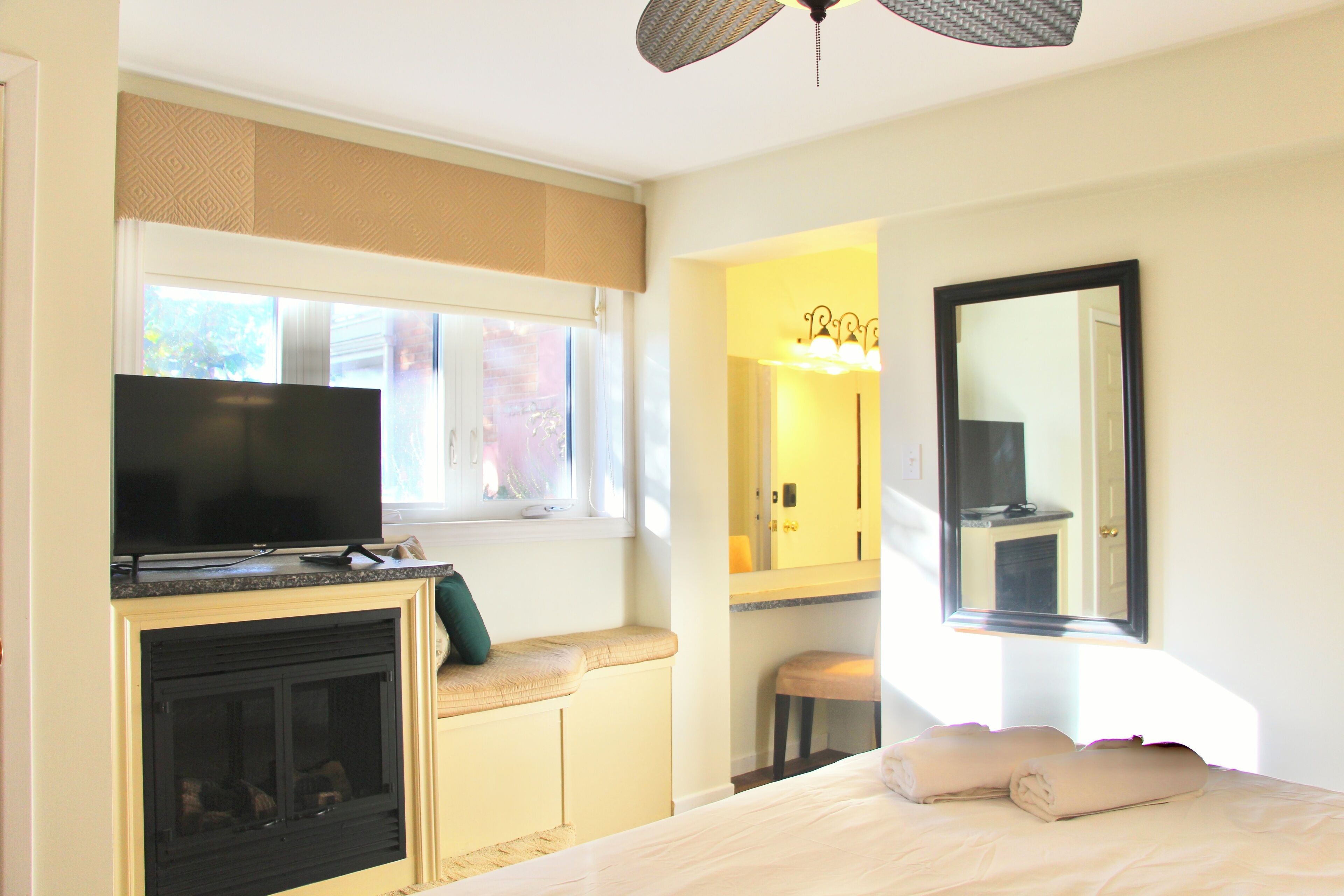 A hotel room with a television on a cabinet, a fireplace, a bed with towels, a window with blinds, and a mirror.