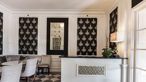 Ein Zimmer mit Schachbrettmuster-Boden, einer Sitzecke mit Tisch und Stühlen, einer dekorativen Wandverkleidung und einem Empfangstresen mit Pflanze.