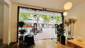 Entrada de hotel con puerta de vidrio, plantas de interior, una mesa de madera y vista a la calle.