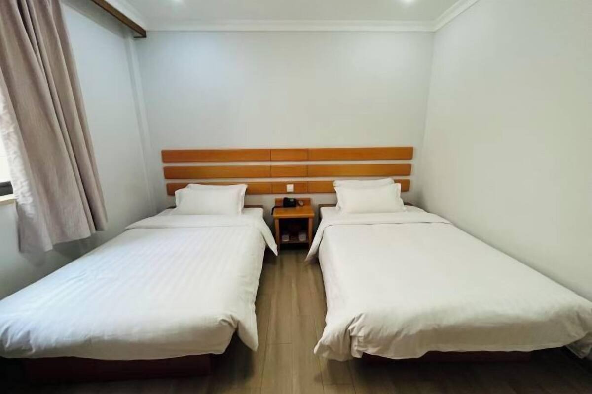 Una habitación con dos camas individuales, un cabecero de madera, una mesita de noche y una ventana con cortinas.