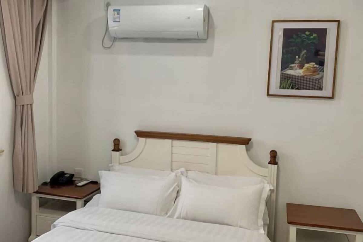 Una habitación de hotel con una cama, una mesita de noche con un teléfono, un cuadro enmarcado en la pared y un equipo de aire acondicionado sobre la cama.
