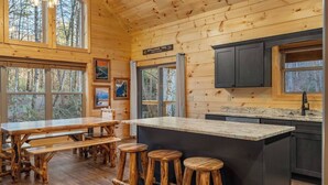 Ein Blockhaus-Interieur mit Holzwänden, einer Küchenzeile, einem Esstisch mit Bänken und einer Bar-ähnlichen Sitzgelegenheit.