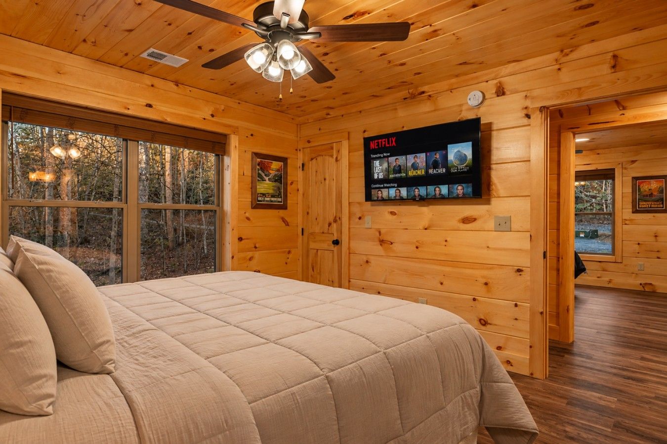 Une chambre dans un chalet en bois, avec un lit, un ventilateur de plafond, un téléviseur fixé au mur diffusant du contenu Netflix et de grandes fenêtres.