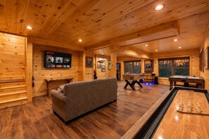 A cozy log cabin interior with a sofa, wooden floors, a foosball table, and a TV mounted on the wall.