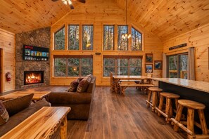 A log cabin interior with a stone fireplace, wooden furniture, and large windows offering a forest view. - Waterfront Swimming + Pet Friendly | Hot tub (Cocke County)