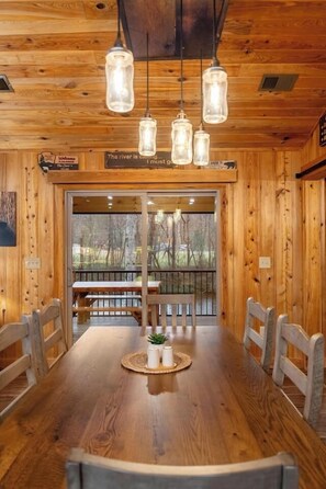 A wooden interior with a long dining table, benches, hanging light fixtures, and a view through a glass door.