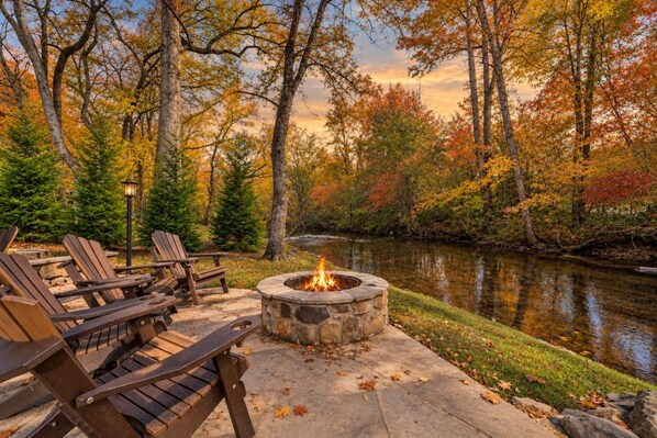 A stone fire pit surrounded by Adirondack chairs on a stone deck, with a river and autumn foliage in the background.