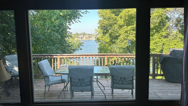 Vue depuis l’intérieur d’une pièce sur une terrasse avec une table et des chaises, donnant sur une étendue d’eau et des arbres.