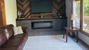 Un salon avec une cheminée, un canapé en cuir marron, une petite table et une vue sur l’extérieur par une fenêtre.