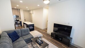Un salon moderne avec un canapé gris, une table basse, un téléviseur à écran plat et un coin cuisine équipé d’un four à micro-ondes et d’un four traditionnel.