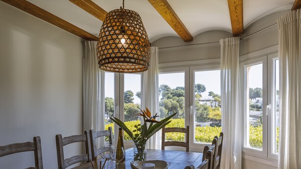 A dining area with a wooden table, chairs, a hanging light fixture, and large windows offering a view of greenery and buildings.