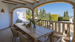 A wooden table with chairs, a vase with flowers, and a fruit bowl on a balcony with a view of the sea and trees.
