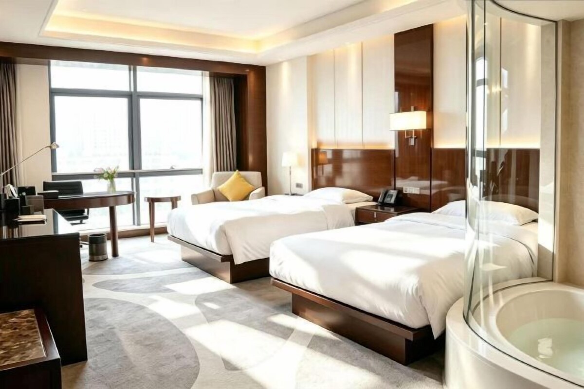 A hotel room with two beds, a desk, a chair, a bathtub, and a large window.