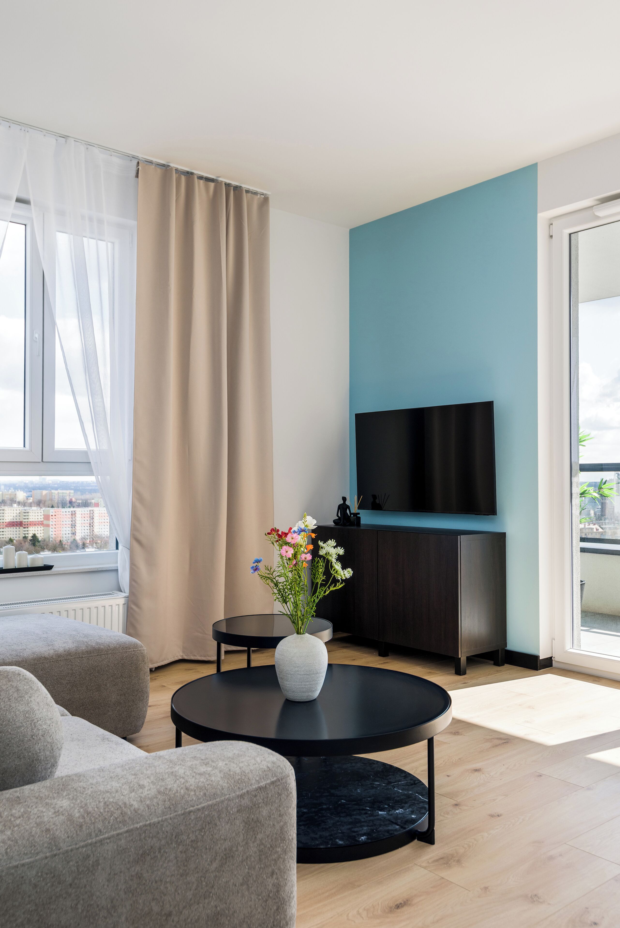 A modern living room with a round black coffee table, grey sofas, a white vase with flowers, a dark wooden cabinet, and a flat-screen TV mounted on a teal wall.