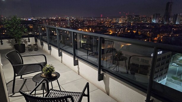 A balcony with a city view at night, featuring a glass railing, two chairs, a small table, and a potted plant. - Sky by Kairos Apartments (Katowice)