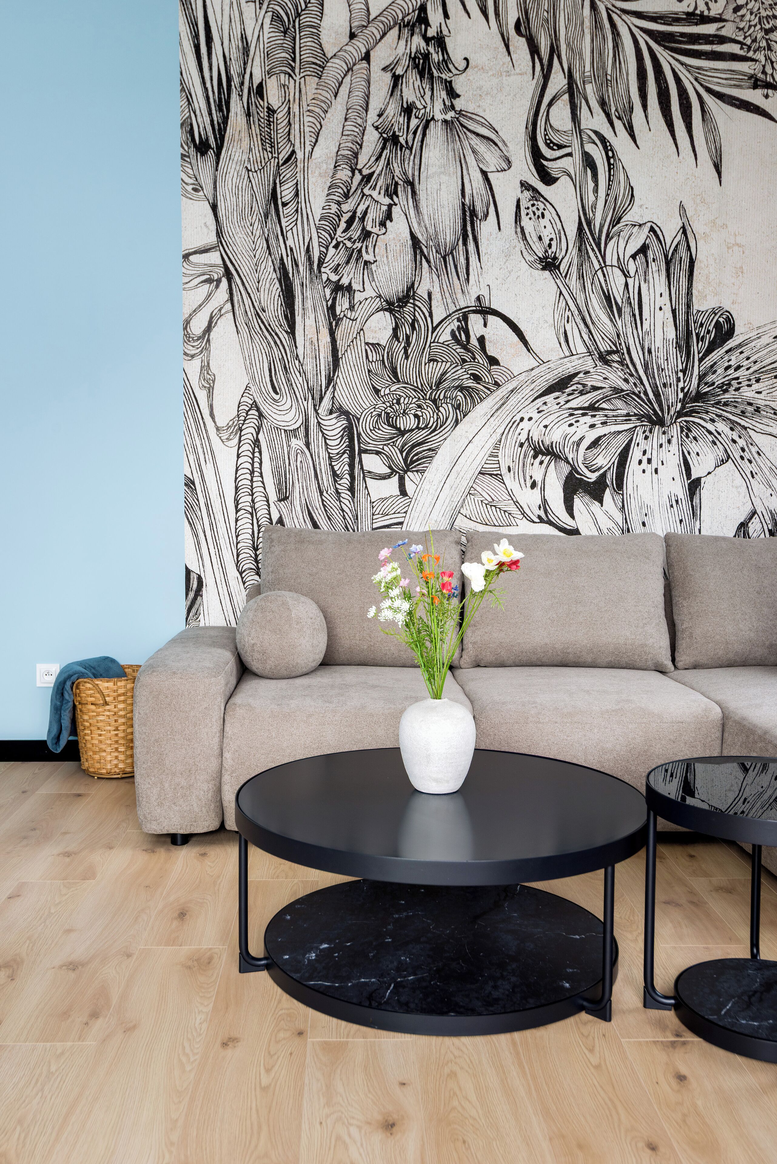 En moderne stue med en grå sofa, et rundt sort sofabord og et stort blomstermotiv i sort og hvidt på væggen.