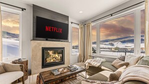 A cozy living room with a fireplace, a large window with a snowy view, and a flat-screen TV mounted above the fireplace. - 4 BR Cozy Upscale Mountain Retreat, Hot Tub, Mountain Views (Crested Butte)
