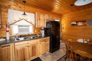 Interior de una cabaña de madera con cocina, que incluye armarios de madera, un refrigerador negro, una mesa de comedor con sillas y una lámpara colgante.