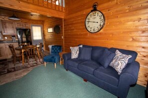 Interior d'una cabana acollidora amb un sofà blau, parets de fusta, zona de menjador i un rellotge de paret gran.