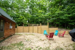 Una cabaña de madera con patio trasero cercado, mobiliario exterior y un bosque denso al fondo.