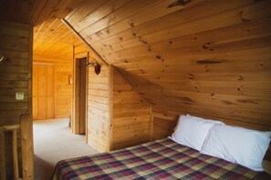 Una habitación de cabaña de madera con techo inclinado, una cama con edredón a cuadros y una puerta visible que da a otra habitación.