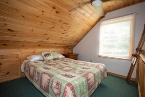 A bedroom with a sloped ceiling, a bed with a patterned bedspread, a nightstand, a window with blinds, and a wooden ladder.