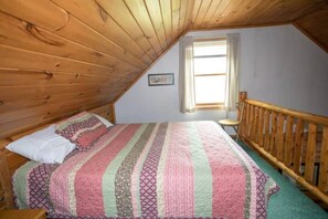 Ein Schlafzimmer mit Holzbalkendecke, einem Bett mit gestreifter Bettdecke, einem Sessel und einem Fenster mit VorhÀngen.
